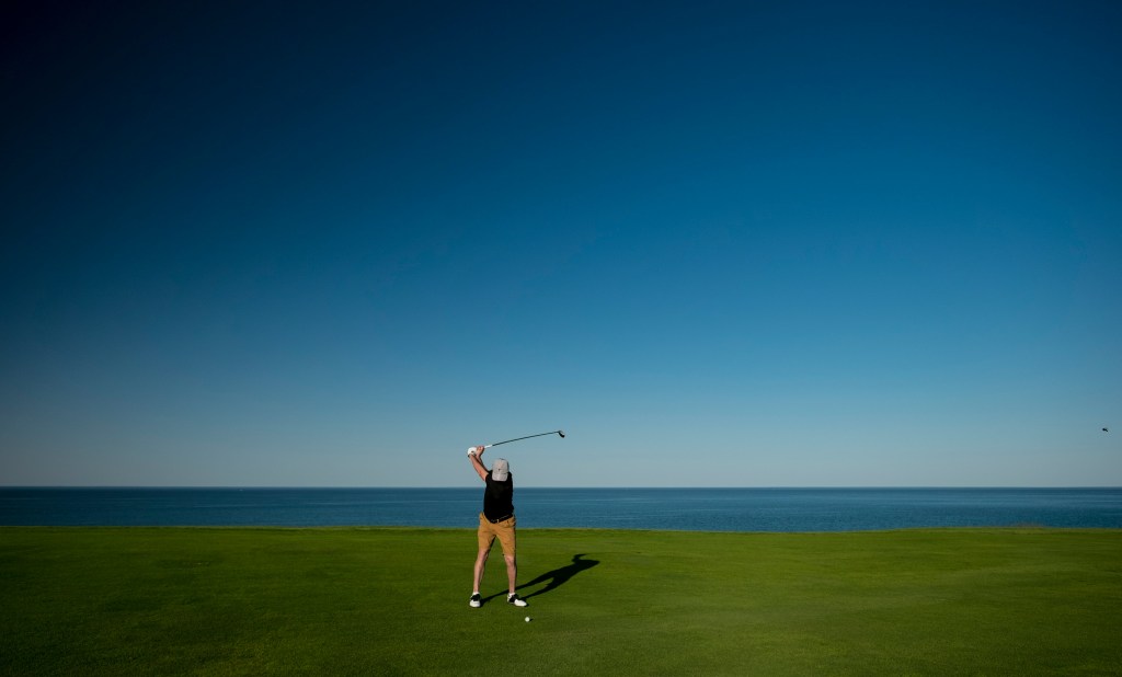 A man in a black shirt and khaki pants takes a golf swing. The perspective is zoomed out to show him as relatively small compared to the vast field he's standing on and the ocean at his back.