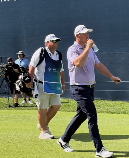 A man in a white hat and pink shirt drinks from a water bottle while a man carrying golf clubs in a white shirt and shorts walks behind him. 